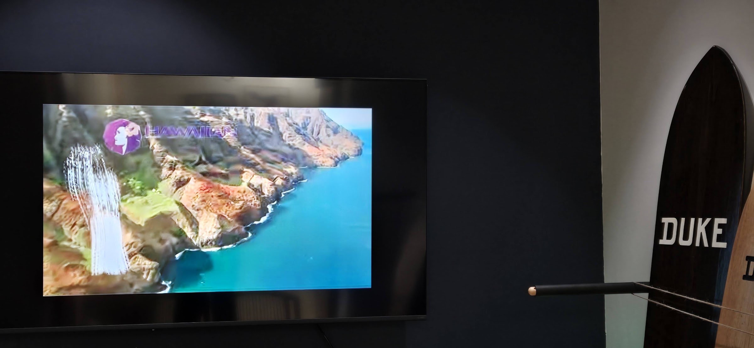A screen on a dark wall showing an aerial view of a Hawaiian coastline, with the edge of a surfboard at the right.